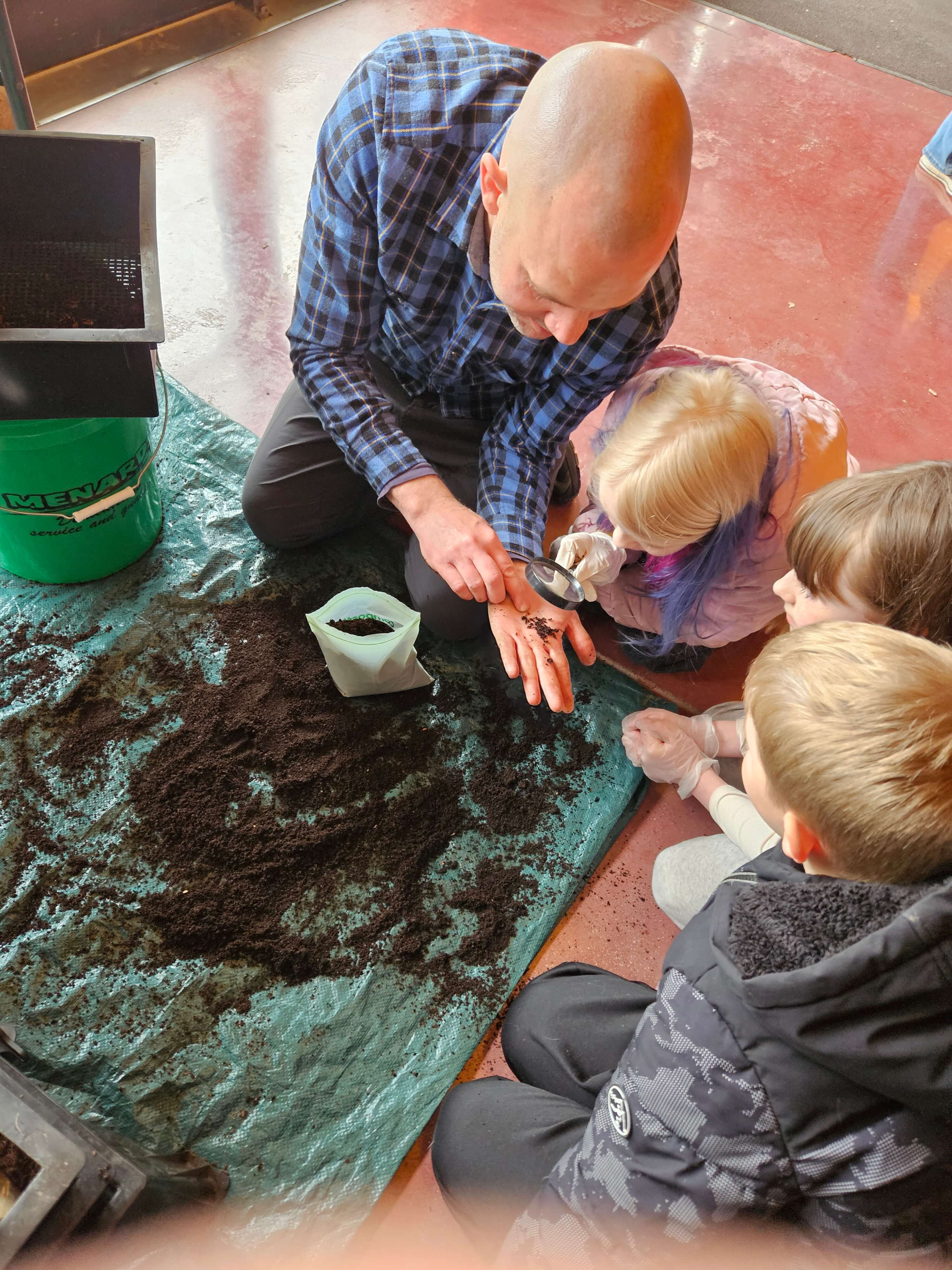 young man holding dirt and worms in his hand to three young children