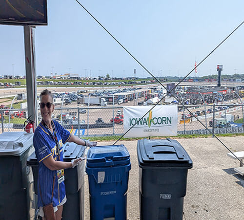 woman in purple and gold jersey standing by a recycling and trash barrel with race track in the background