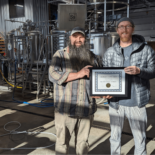 Neil Blair, head brewer. and Elliot Thompson, owner of Alluvial Brewing stand on the floor of their brewery holding their gold certification plaque