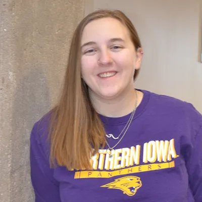 Young woman wearing a University of Northern Iowa purple shirt leaning against a cement pillar