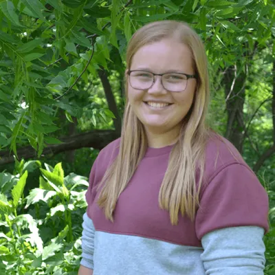 Graphic design intern Haley Christoffer stands before a tree with healthy green leaves.